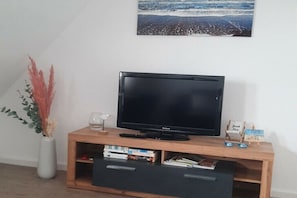 A television on a wooden stand with a beach-themed picture above it, a vase with dried plants, and a small wooden shelf with items. - Holiday apartment with 2 bedrooms (Borkum)