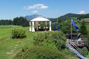 Ein Gartenpavillon mit weißem Baldachin und weißen Vorhängen, umgeben von viel Grün und einem kleinen Gebäude mit einer blauen Flagge.