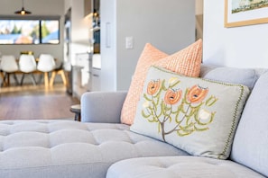 Ein modernes Wohnzimmer mit einem grauen Sofa, einem Zierkissen mit floraler Stickerei und einem Kissen mit Schachmuster.