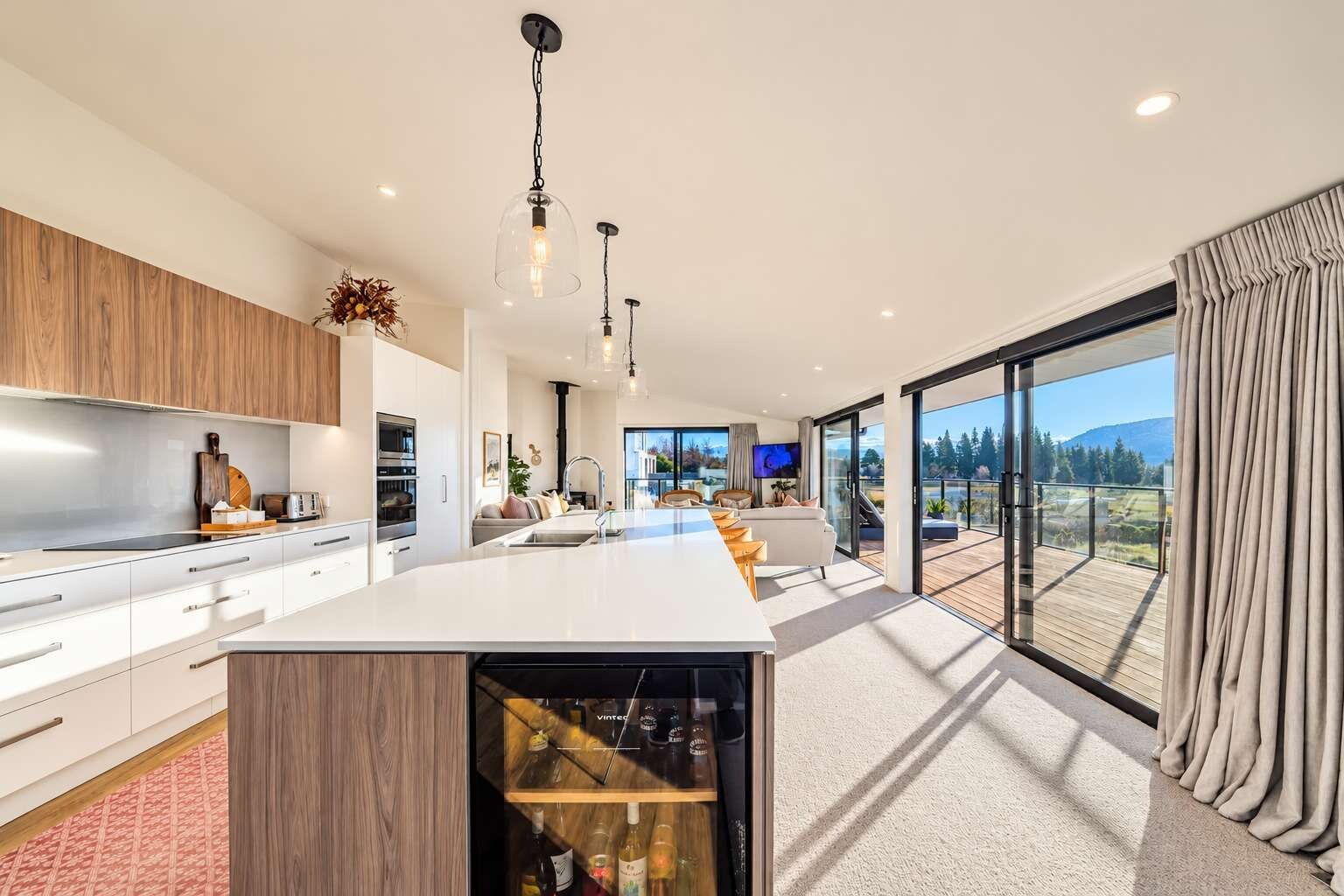 A modern kitchen with a central island, a dining area, and a large sliding glass door leading to a deck with a view of the outdoors.