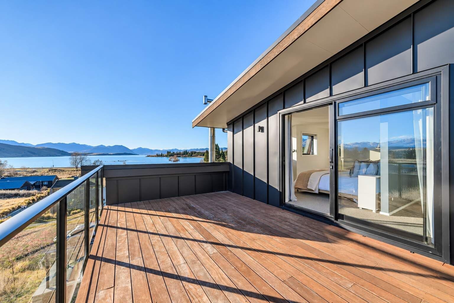 Une chambre d’hôtel moderne dotée d’une grande porte vitrée, d’une terrasse en bois et offrant une vue sur un lac et des montagnes.
