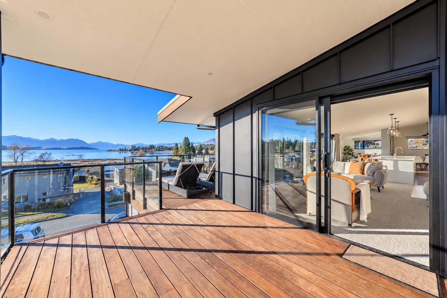 Un balcon moderne avec une terrasse en bois, des portes vitrées donnant sur un espace intérieur et une vue sur un lac et des montagnes.