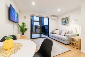 A modern living room with a sofa, a dining table with chairs, a TV mounted on the wall, and a sliding glass door leading to a balcony. - Modern 2BR with Patio | Christchurch CBD (Christchurch)