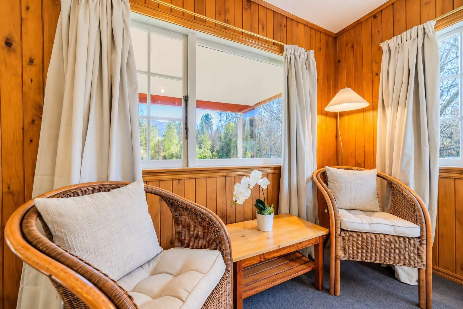 A cozy room with wooden paneled walls, two wicker chairs with cushions, a small table, and a window with curtains.