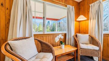 A cozy room with wooden paneled walls, two wicker chairs with cushions, a small table, and a window with curtains.