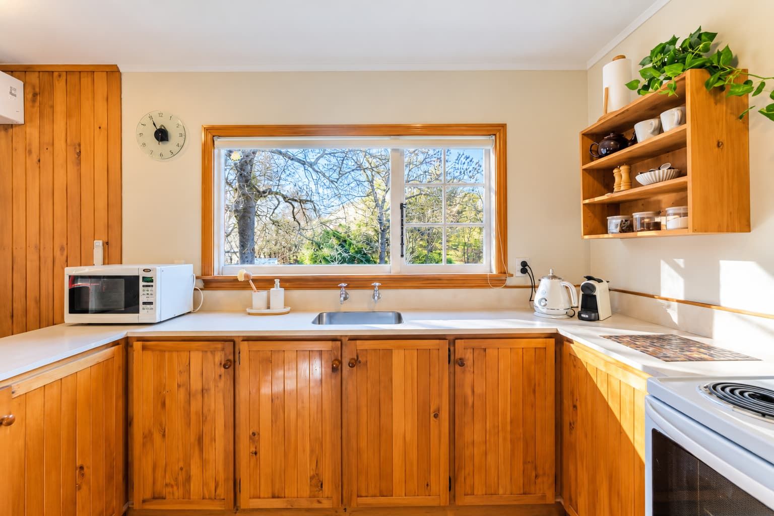 A kitchen with wooden cabinets, a microwave, a sink, and a window with a view of trees.