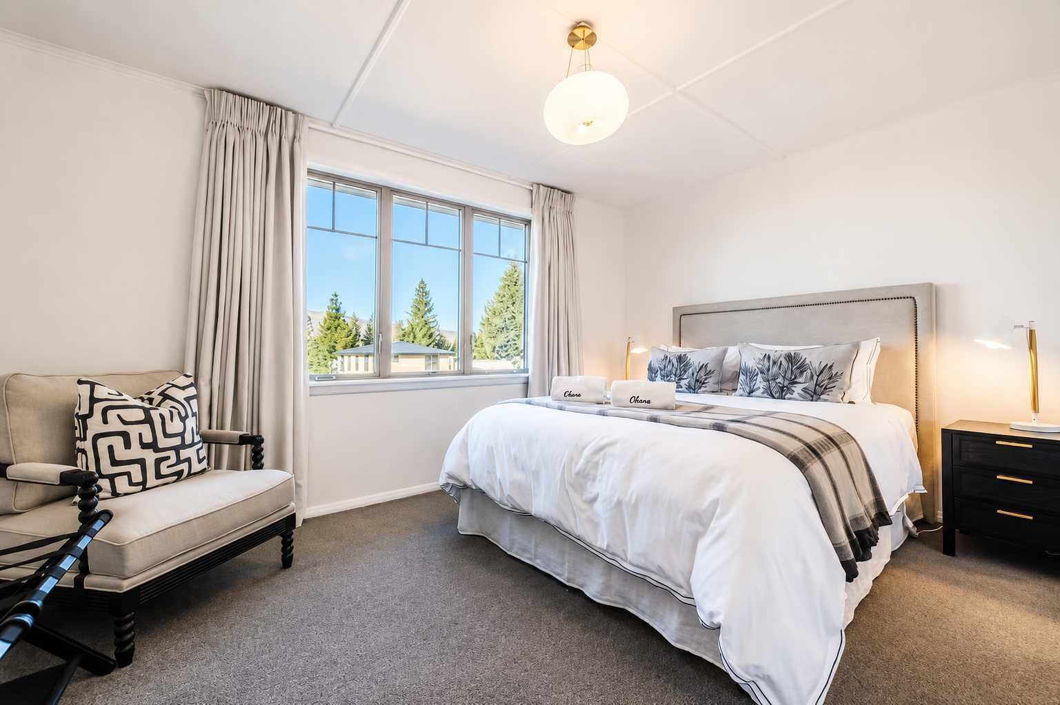 A hotel bedroom with a large bed, a chair, a nightstand, and a window with a view of trees.