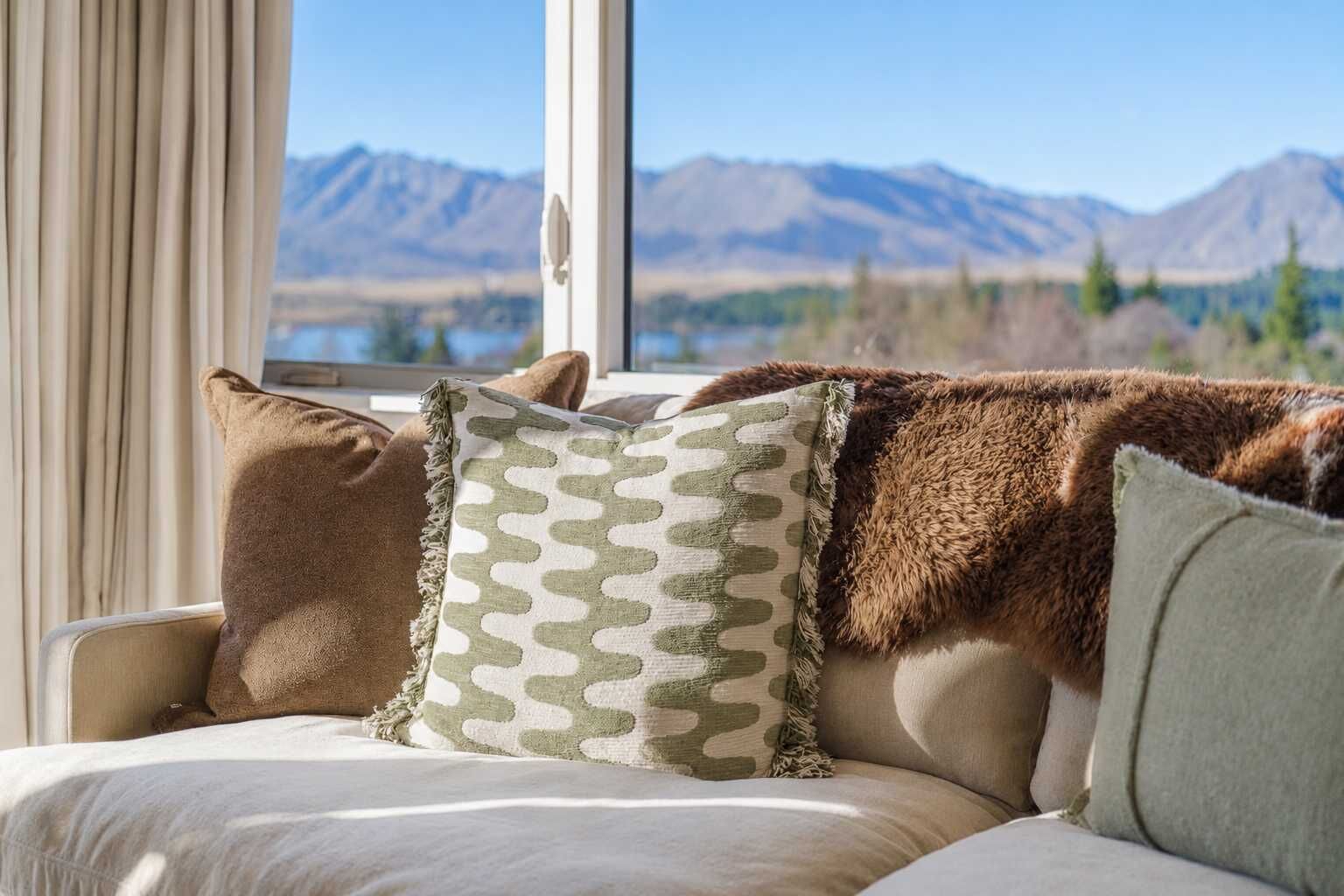 A room with a sofa and pillows, a window with a mountain view, and a curtain.