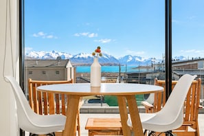 A round table with white chairs and a vase with flowers, overlooking a mountainous landscape with a body of water.