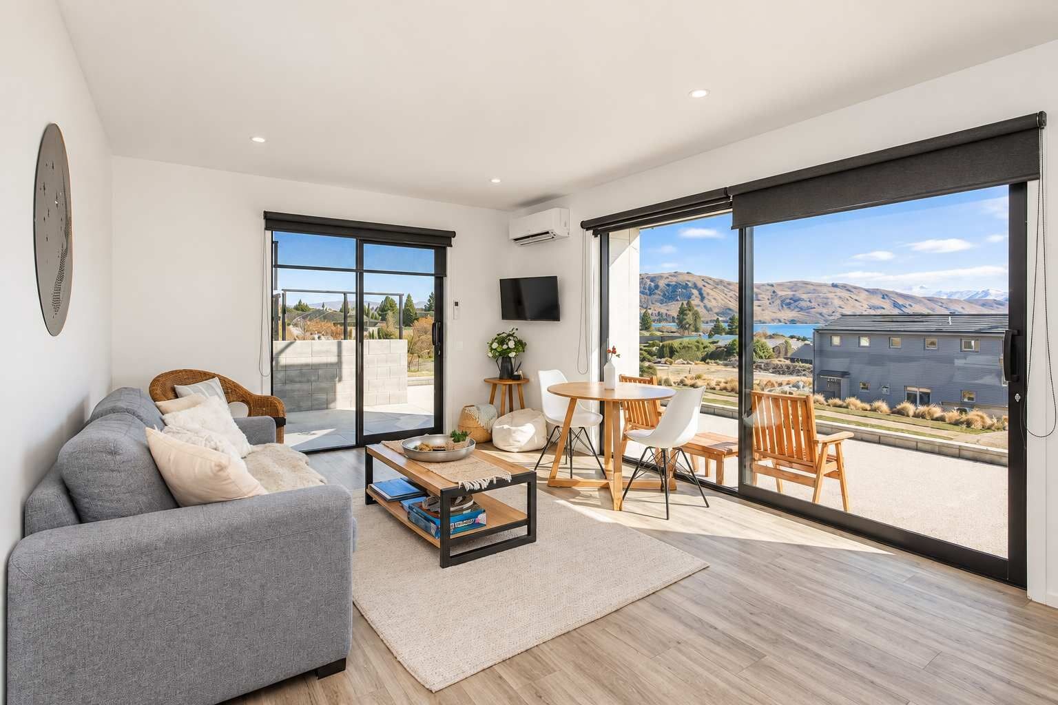 A modern living room with a grey sofa, a coffee table, and a dining area with chairs and a table. Large sliding glass doors lead to a balcony with a view of a building and landscape.
