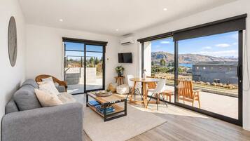 A modern living room with a grey sofa, a coffee table, and a dining area with chairs and a table. Large sliding glass doors lead to a balcony with a view of a building and landscape.