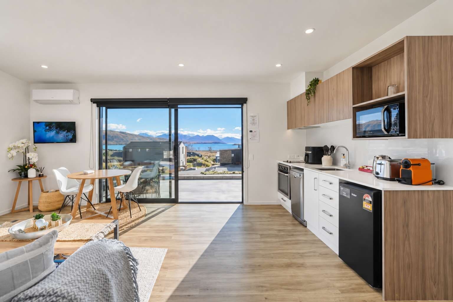 A modern kitchen with a view of mountains, a dining area with a table and chairs, and a living space with a sofa and a TV.