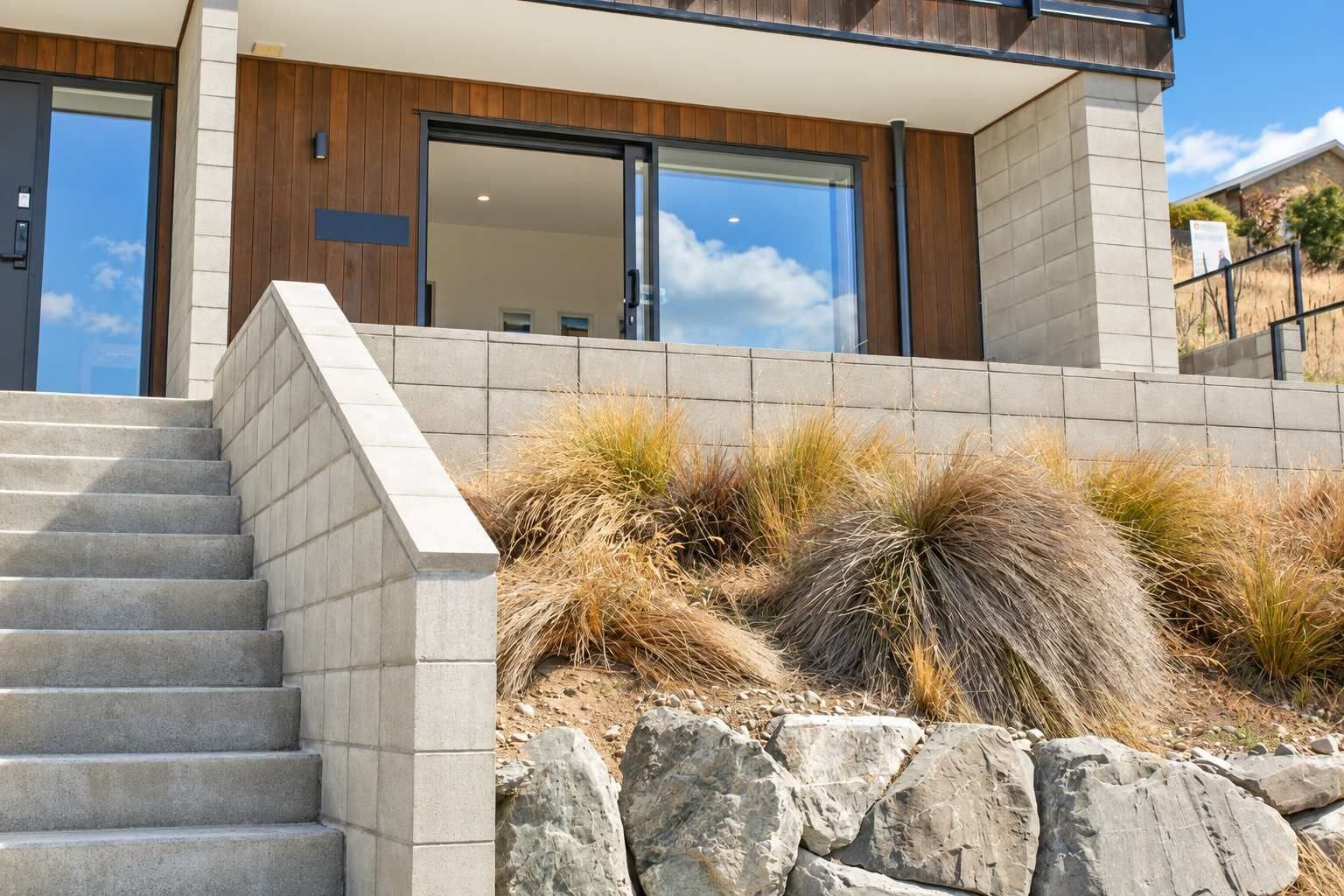 Un bâtiment moderne avec un escalier en pierre menant à une porte en bois, entouré d’herbe sèche et de rochers.