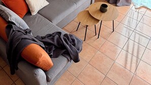 Un salon moderne avec un canapé gris, des coussins orange et blancs, une table basse ronde, un téléviseur à écran plat, une décoration murale en forme de boussole et une fenêtre avec des rideaux.