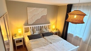 Une chambre avec un grand lit, deux tables de chevet avec des lampes, une œuvre d’art accrochée au mur, un panier en osier et un luminaire au plafond.