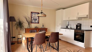 Une cuisine moderne avec des meubles blancs, une table à manger entourée de quatre chaises, un luminaire suspendu et une plante en pot.
