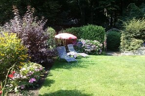 Ein Garten mit Rasen, zwei gestreifte Liegestühle, einem Tisch und einem roten Sonnenschirm.