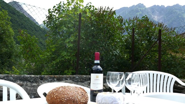 Uma mesa com um pão, uma garrafa de vinho e uma faca em uma tábua de corte, com montanhas ao fundo.