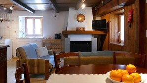 Uma sala de estar acolhedora com lareira, uma mesa de jantar com uma tigela de laranjas e uma televisão fixada na parede.