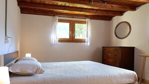 Quarto com teto de madeira, cama com colcha branca, cômoda, espelho e janela com cortinas.