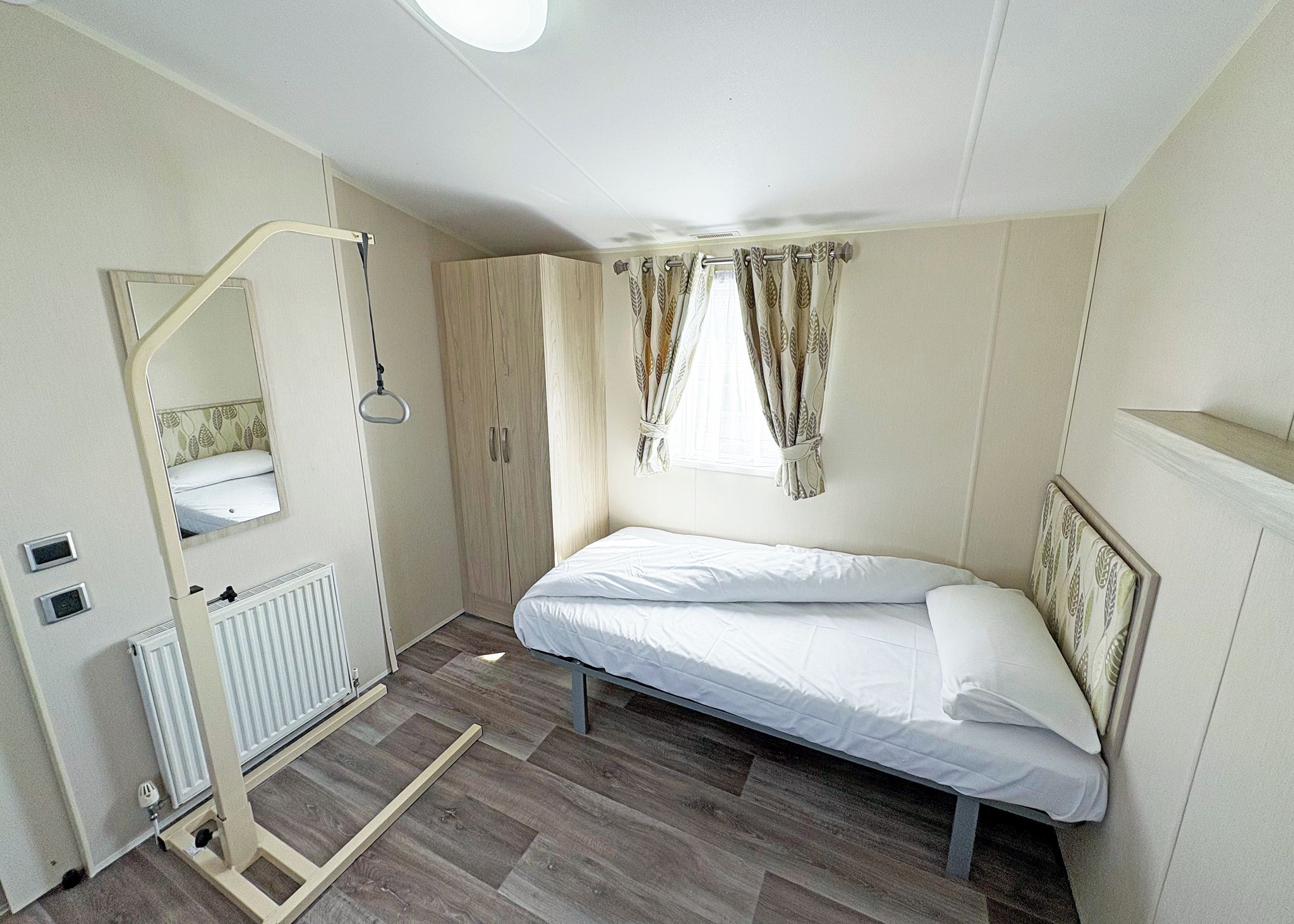 Een kleine, goed verlichte kamer met een bed, een radiator, een spiegel en een kledingkast.