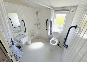 Ein kleines Badezimmer in Weiß mit Toilette, Waschbecken und Dusche. In der Nähe von Toilette und Dusche befinden sich Haltegriffe, und es gibt ein Fenster mit Blick.