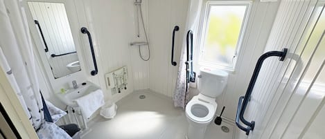 A small, white bathroom with a toilet, sink, and shower. There are grab bars near the toilet and shower, and a window with a view.