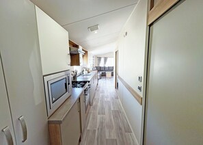 A narrow kitchen with a microwave, stove, and sink. A small dining area with a table and chairs is visible in the background. - Purn Gold 6 WF (Pet) (Bleadon, Weston-Super-Mare)