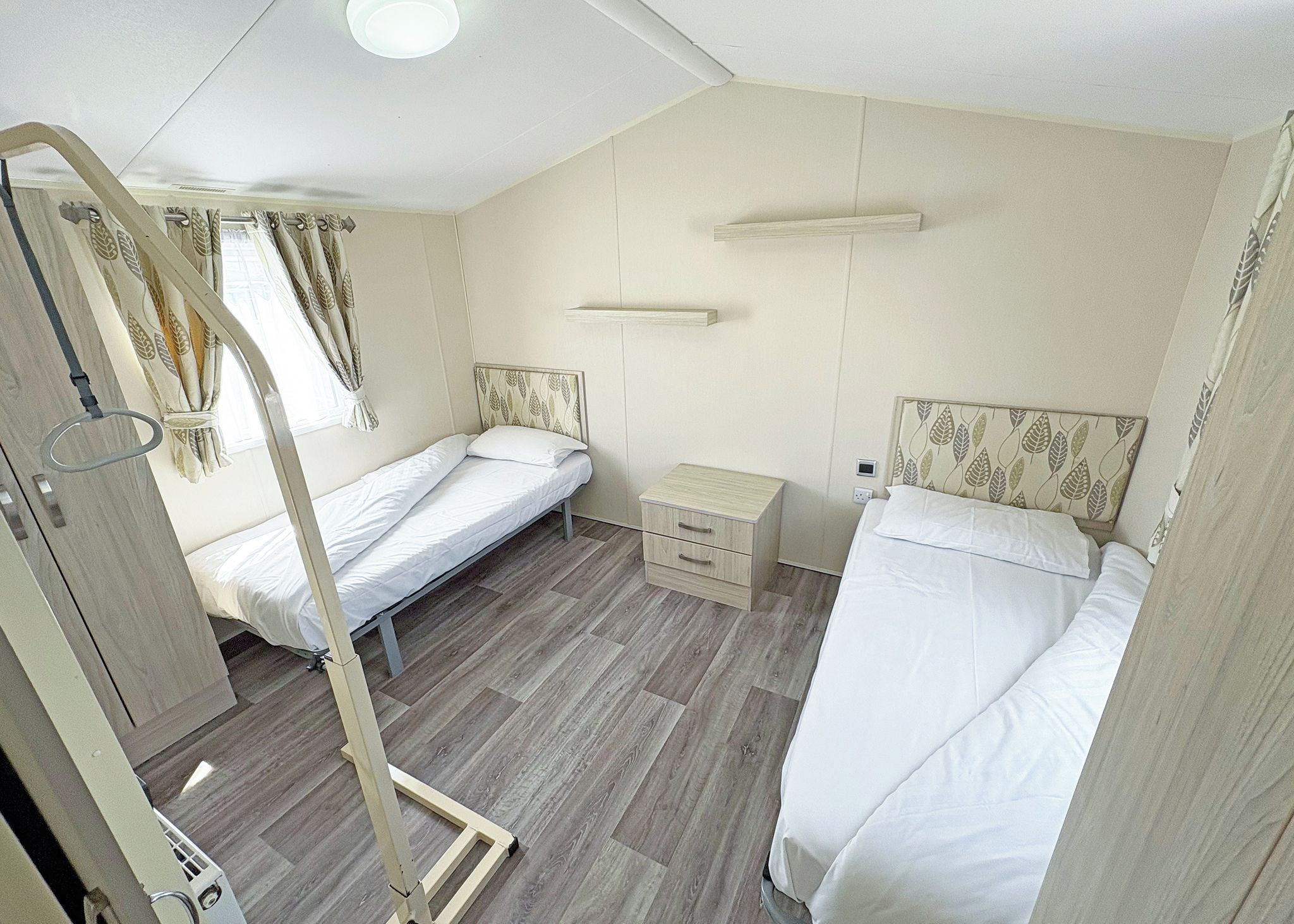 A compact room with two beds, a bedside table, a small cabinet, and a curtain with a leaf pattern.