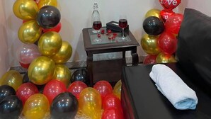 A room with a red 'love' sign on the wall, heart-shaped balloons, a small table with a bottle and glasses, and a black sofa with a white towel.
