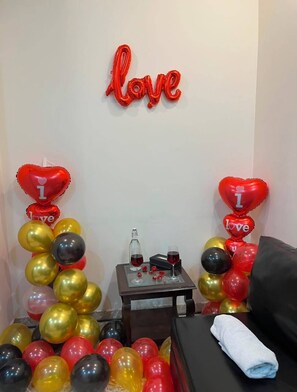A room with a red 'love' sign on the wall, heart-shaped balloons, a small table with a bottle and glasses, and a black sofa with a white towel.