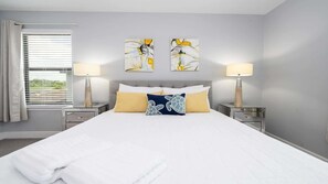 A hotel room with a large bed, two bedside lamps, artwork on the wall, and a window with blinds.