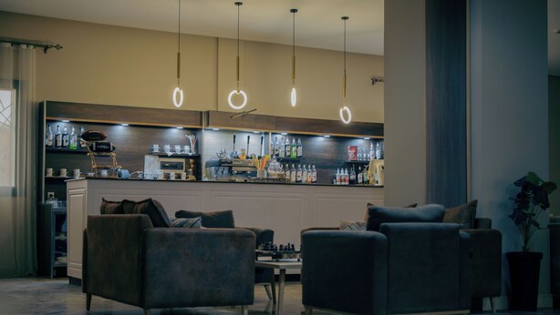 A modern bar area with a counter, shelves of bottles, and hanging lights. There are two sofas in the foreground. - Hotel Asslouni (Djelfa)