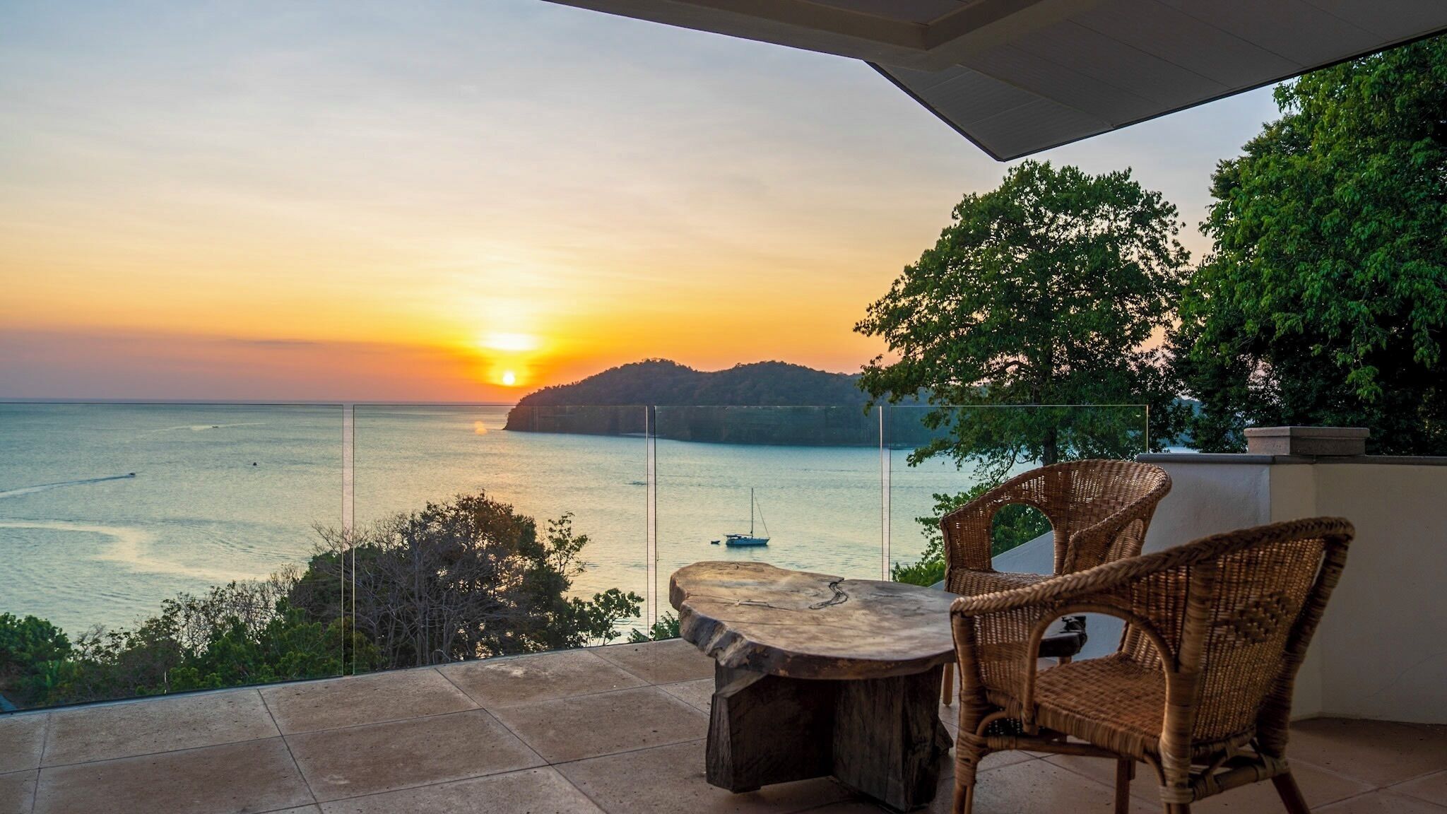 A balcony with a table and chairs, overlooking a sunset over the sea.