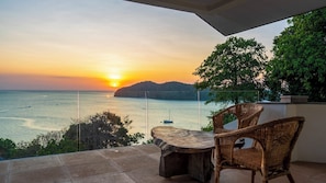 Un balcon avec une table et des chaises, d’où l’on peut admirer le coucher de soleil sur la mer.