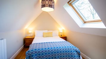 Un dormitorio con techo inclinado, un tragaluz, una cama con colcha estampada, dos lámparas de noche y una mesita de noche.