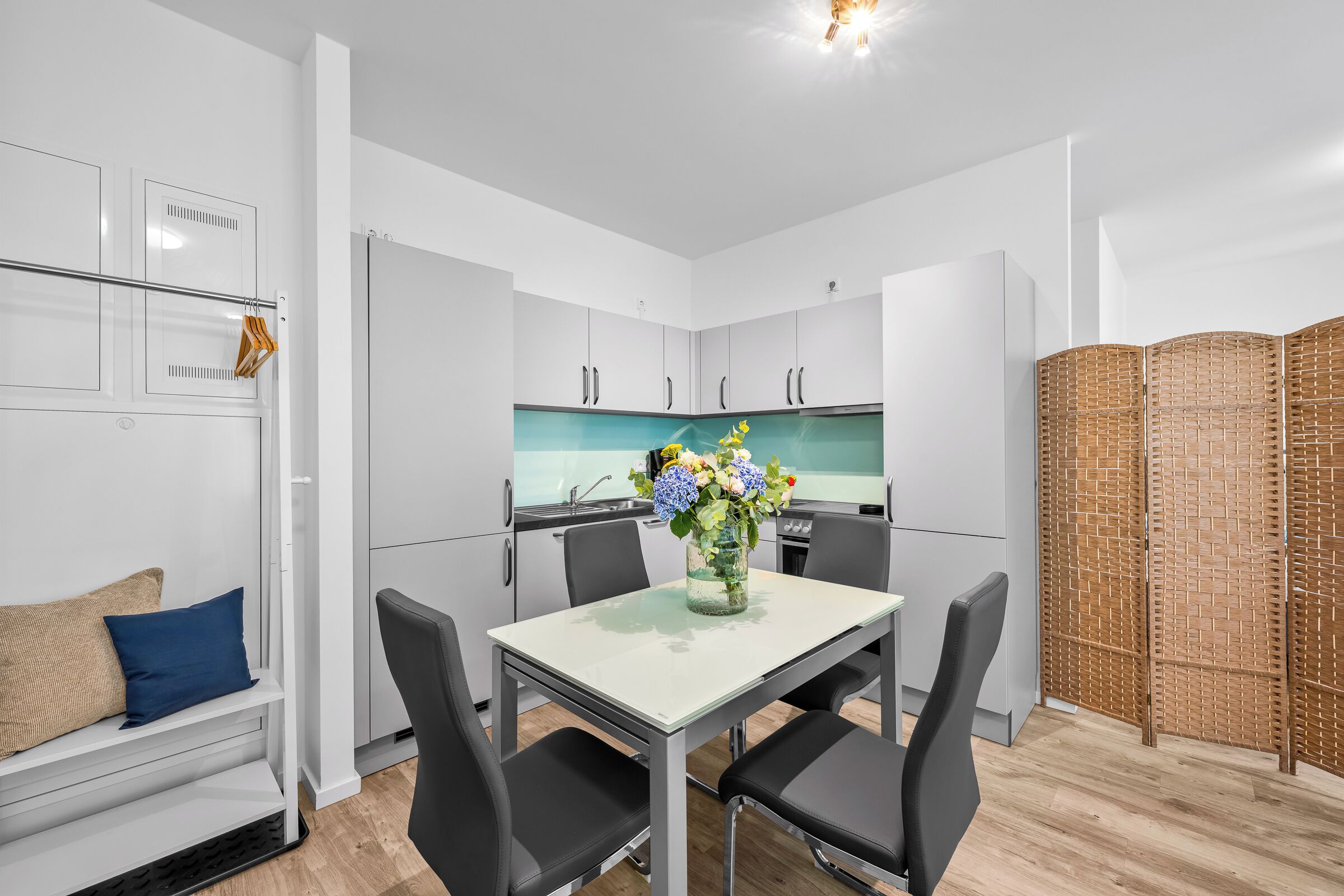 Une cuisine moderne avec des meubles blancs, une table à manger avec des chaises, un vase de fleurs et une cloison en bois.