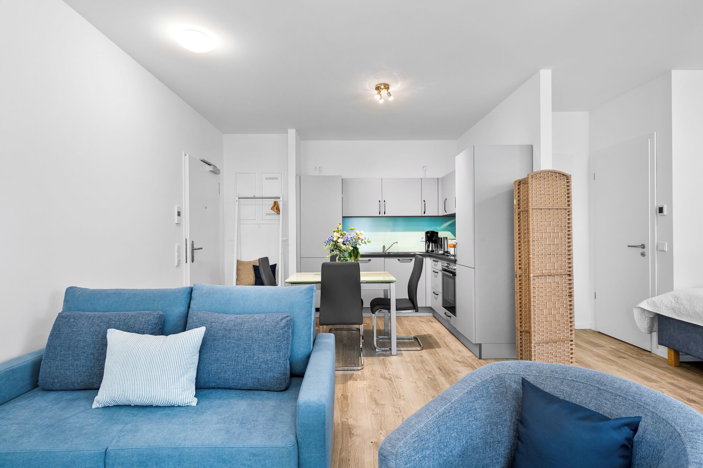Una sala de estar moderna con sofá y sillón, un comedor con mesa y sillas, y una cocina con alacenas blancas y una pared de ladrillo decorativa.