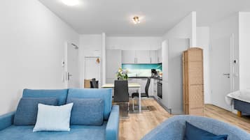Una sala de estar moderna con sofá y sillón, un comedor con mesa y sillas, y una cocina con alacenas blancas y una pared de ladrillo decorativa.