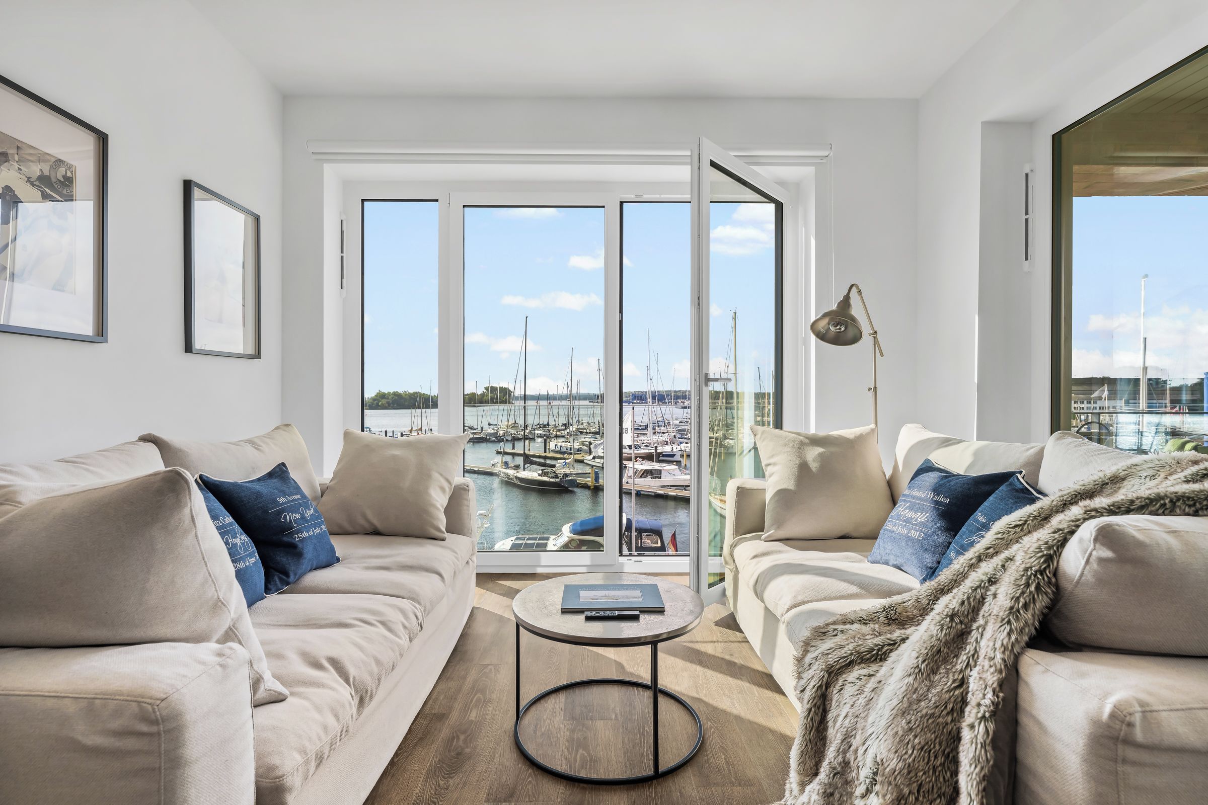 A modern living room with a sofa set, a coffee table, and a view of a marina through large windows.