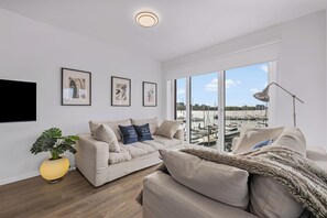 A modern living room with a sofa, a large window with a marina view, and framed artwork on the wall. - Modern Apartment Pure Travemünde with Terrace, Parking and Partial Sea View (Lübeck)