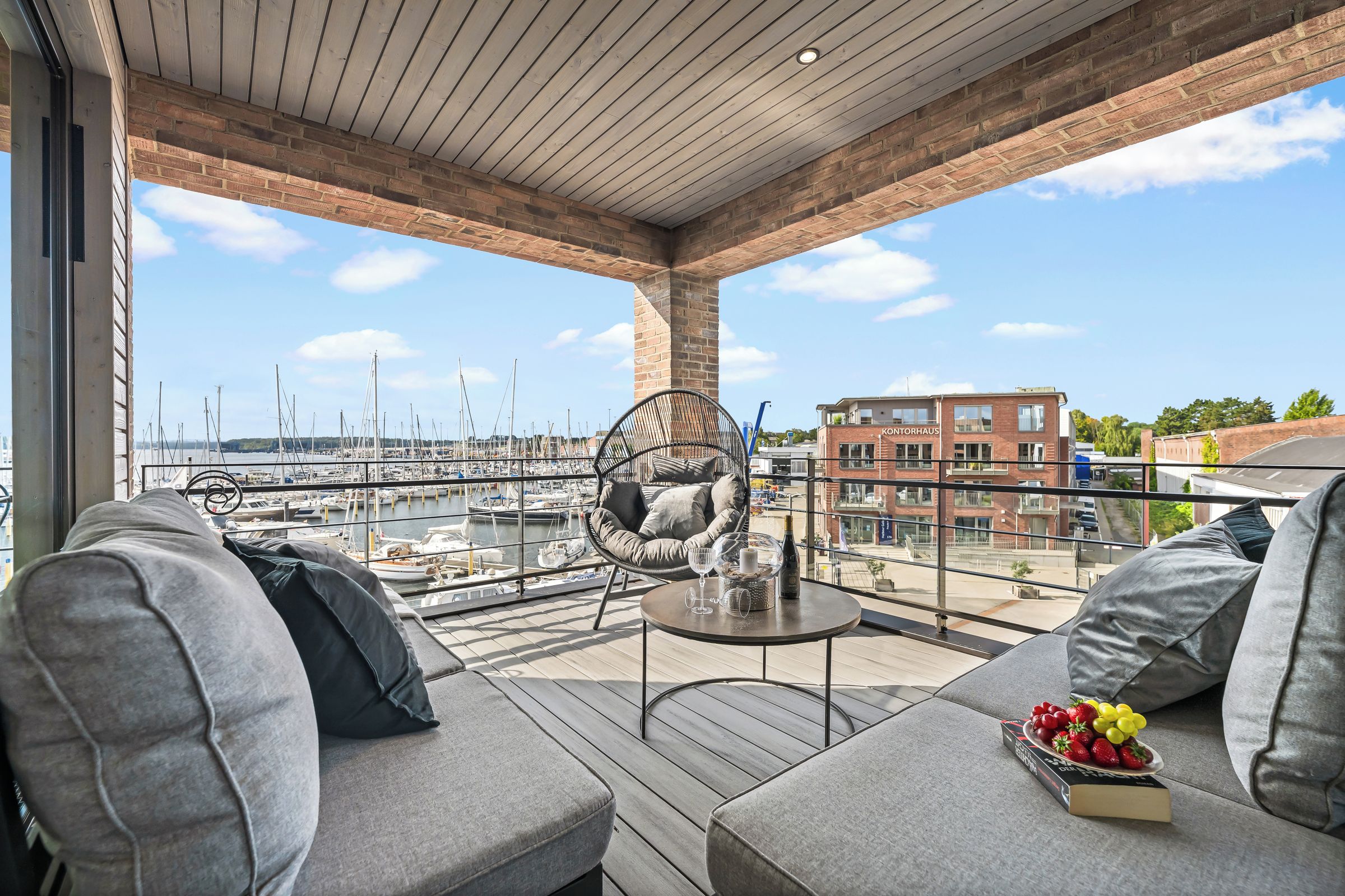 Un balcon donnant sur une marina, une table ronde avec une bouteille et des verres, et un canapé avec des coussins.
