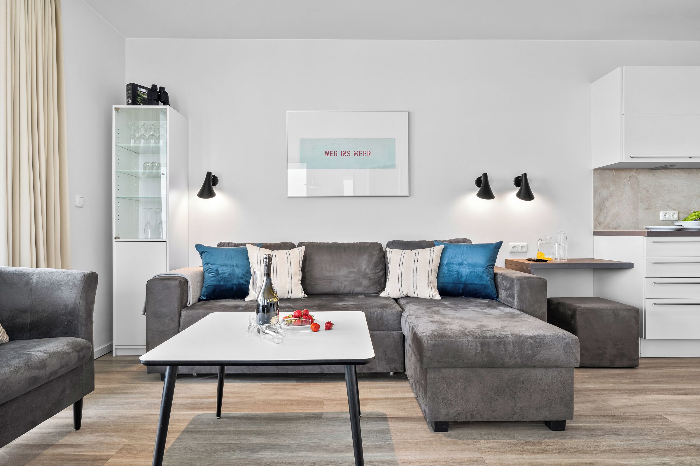 A modern living room with a grey sofa, a white coffee table, and a kitchen area in the background.