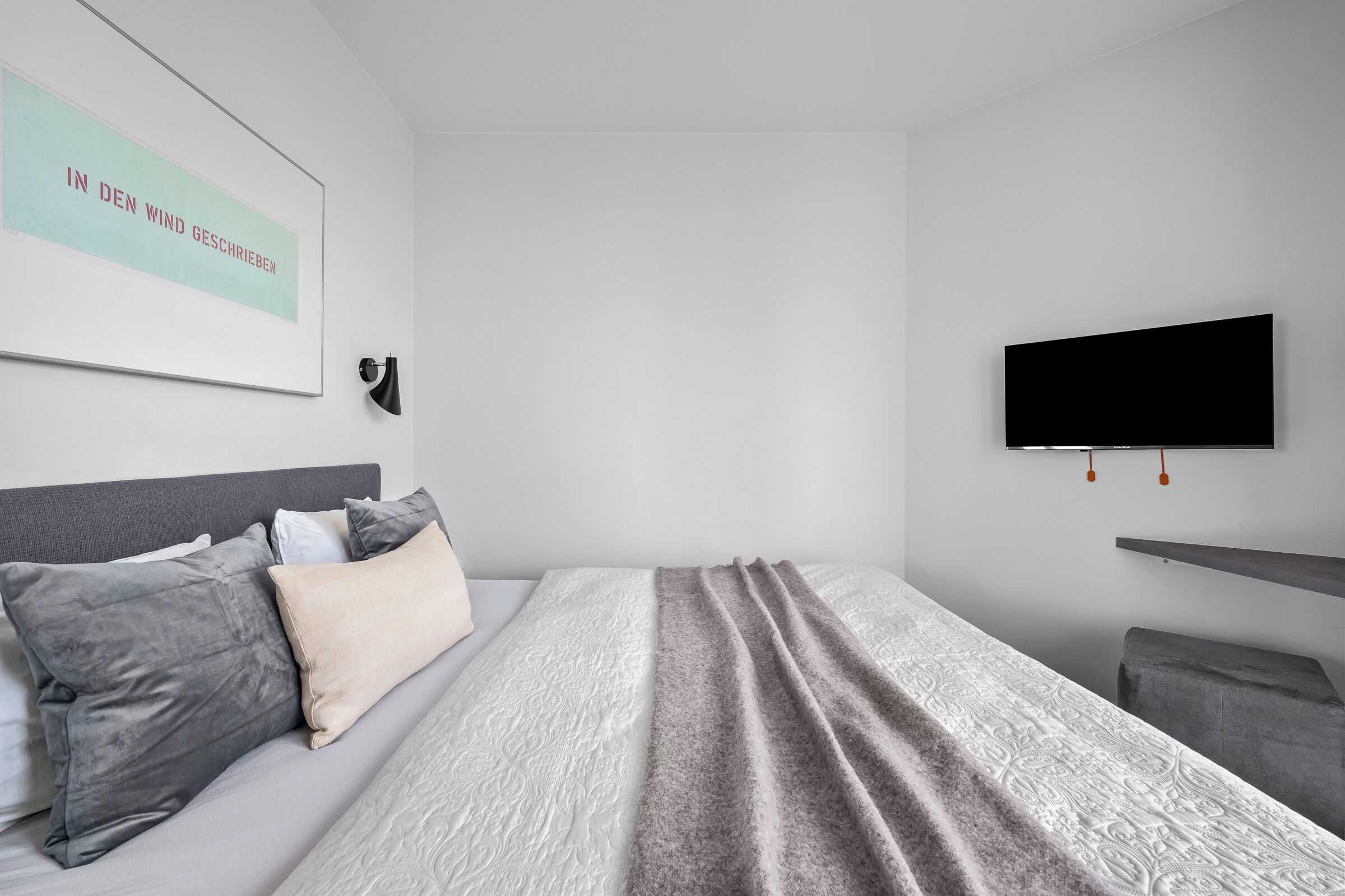 Ein modernes Schlafzimmer mit einem Bett, Kissen, einem an der Wand montierten Fernseher und einem Wandregal.