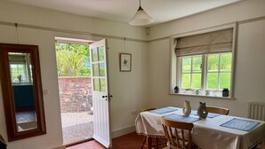 A dining area with a table set for two, a wooden chair, a white door, and a window with a view of greenery. - Cosy Lodge House set in the majestic Quantock Hills (Bishops Lydeard)