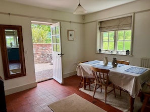 Un coin repas avec une table dressée pour deux personnes, une chaise en bois, une porte blanche et une fenêtre donnant sur un paysage verdoyant.