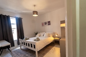 A bedroom with a bed, bedside table, lamp, and a view of the outside.