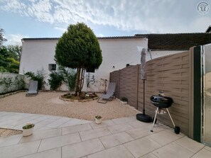 A patio with a white building, a tree, a barbecue grill, and two lounge chairs.