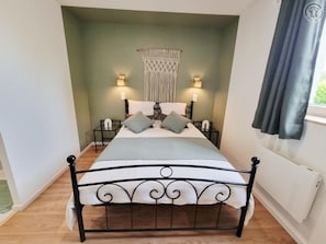 Una cama con estructura metálica, dos mesitas de noche con lámparas, un colgador de macramé en la pared y una ventana con cortinas.