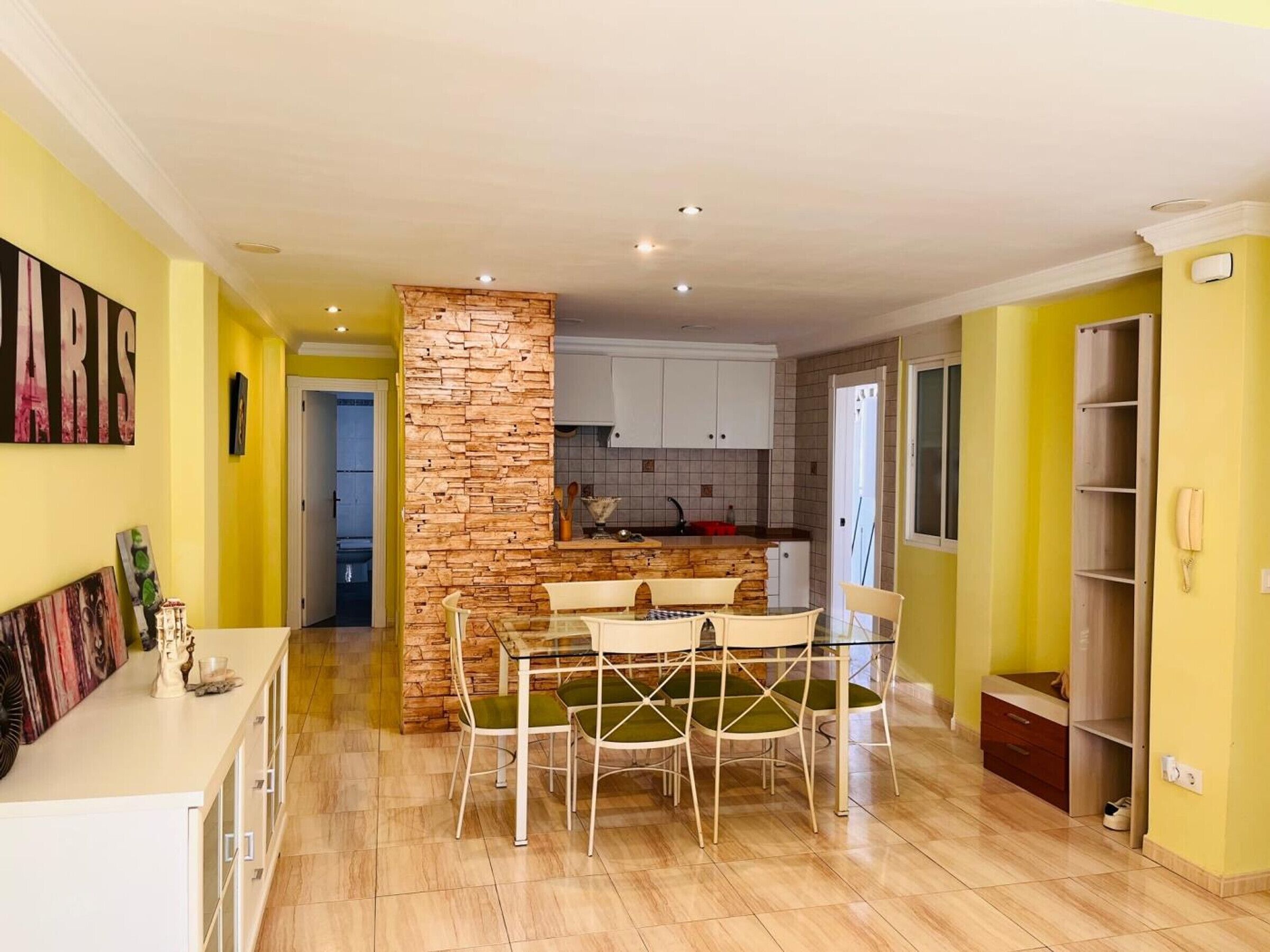 Une cuisine moderne avec un mur d’accent en pierre, un coin repas avec une table et des chaises en verre, et un mur jaune orné d’un téléphone.